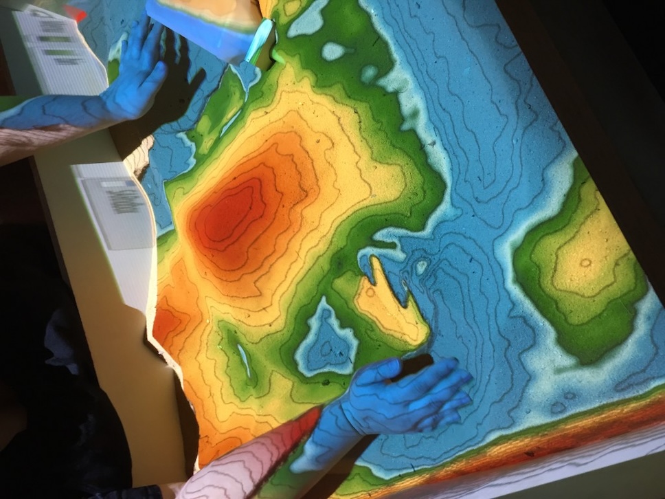 Decorative photo of the GIS Sandbox in use: a projector displays bands of colored light on top of a topographic formation of sand to approximate a topographic map in 3D