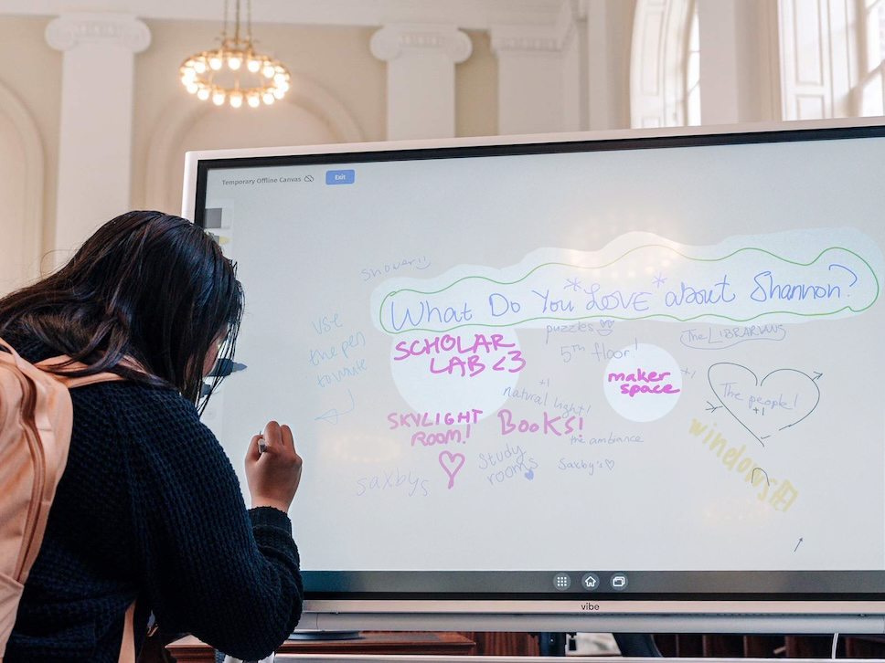 Decorative photo of a student writing on a monitor displaying other students' answers to 'What do you love about Shannon (Library building)?', where previous students have written 'Scholar Lab <3' and 