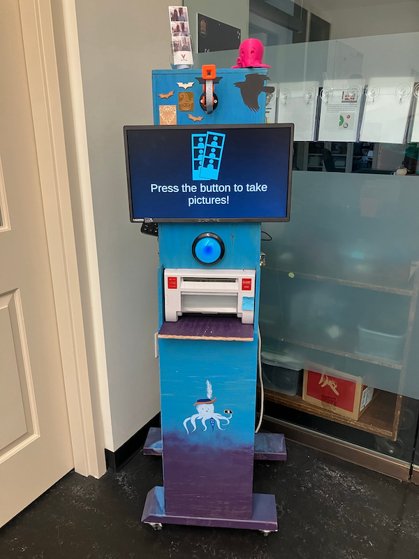 Photo of the Scholars' Lab Makerspace Photobooth, a painted stand with monitor, large button, and camera that takes then prints photobooth-style photo strips
