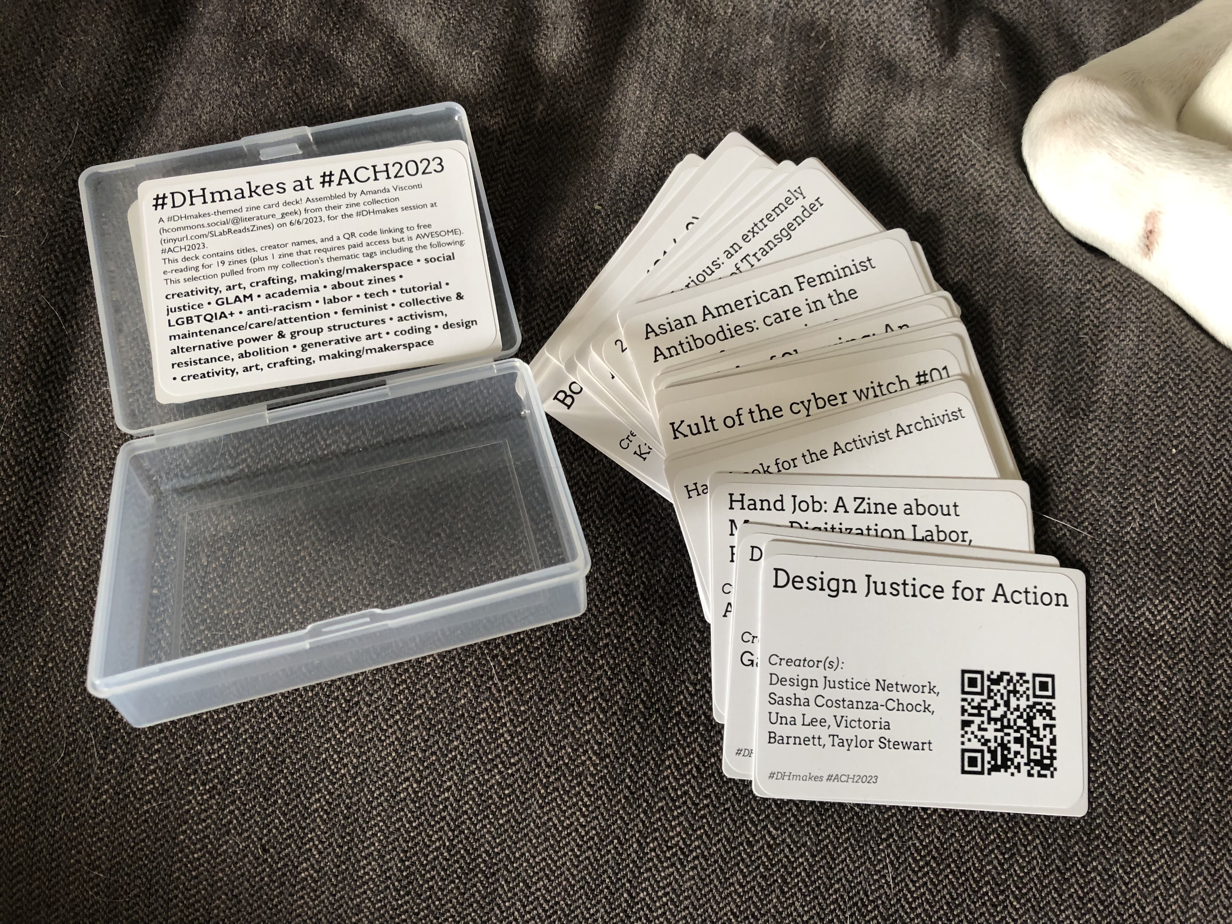 Photo of an open plastic card deck case, next to a hand of playing-card size cards spread out to show the cards each display the title and authors of a zine plus a QR code that links to a digital copy of the zine for reading.