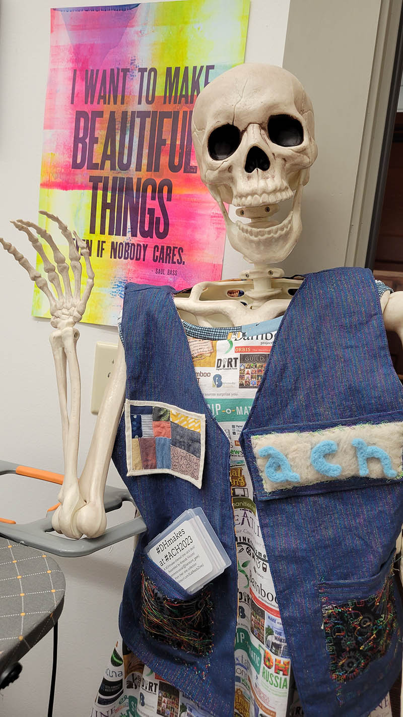 Photo of a full-size skeleton model, Quinn Dombrowski's "Dr. Cheese Bones", with one hand up waving, wearing a denim vest decorated with various small crafting projects made by multiple members of the #DHmakes community including a felted "ACH" patch and a tiny data visualization quilt patch